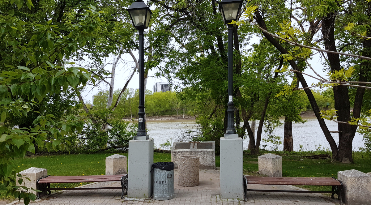 two wooden backless benches with metal legs on opposite sides of two Victorianesque lamp posts in front of several trees along the bank of a river in the background