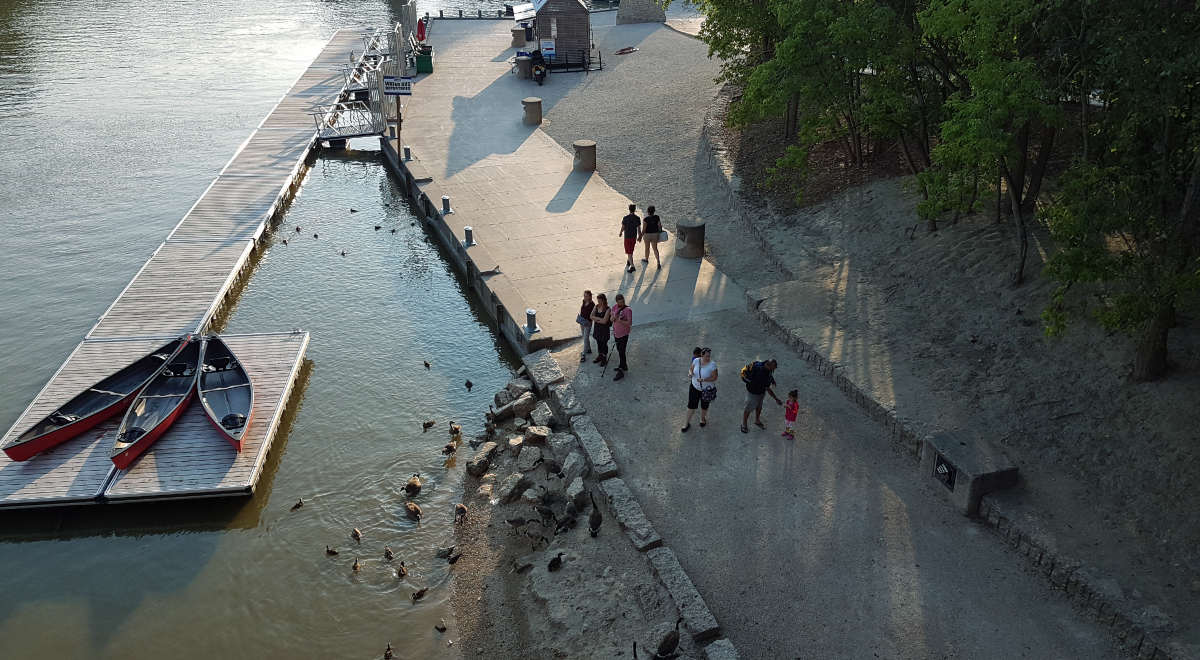 a shot from above of a makeshift dock on a river with three canoes on one side, and a walkway along the river bank with several people standing around