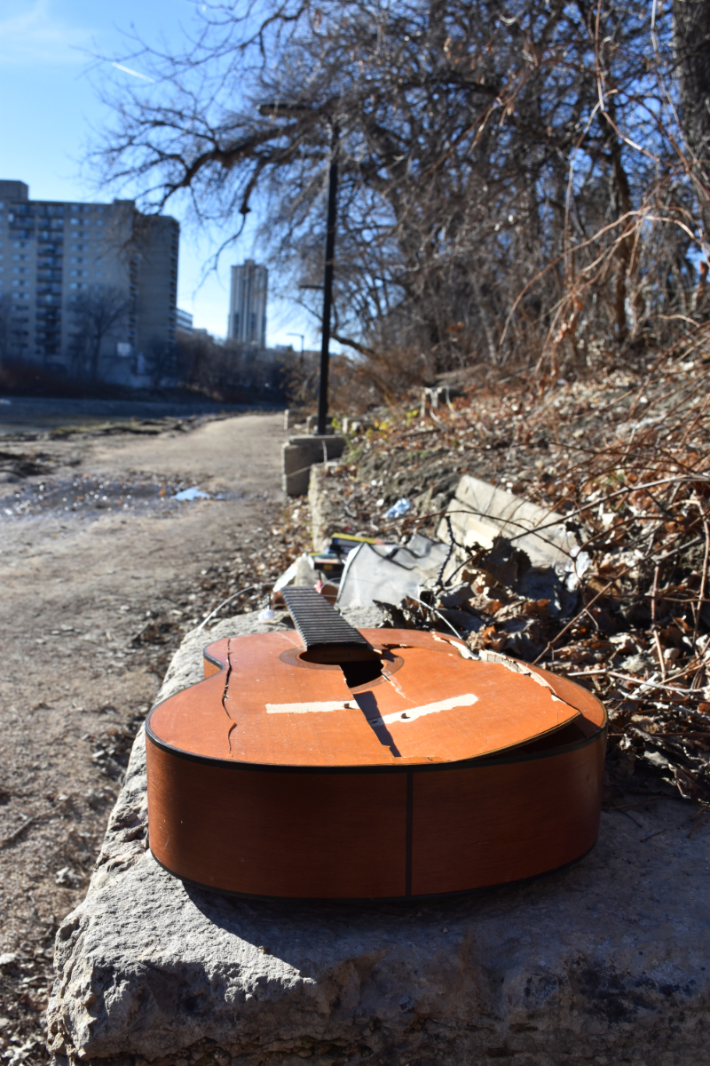 a broken acoustic guitar lying on a stone platform next to a wooden bench along a gravel path next to a river